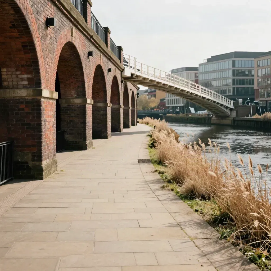 Leeds riverside towpath with brick arches, footbridge shadow, and office blocks in warm midday sun.